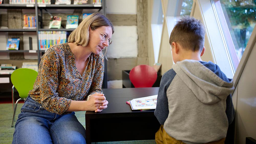 Eine Lehrperson sitzt an einem kleinen schwarzen Tisch und liest mit einem Jungen ein Buch. Sie befinden sich in der Bibliothek. Die Fenster sind rhombusförmig. 