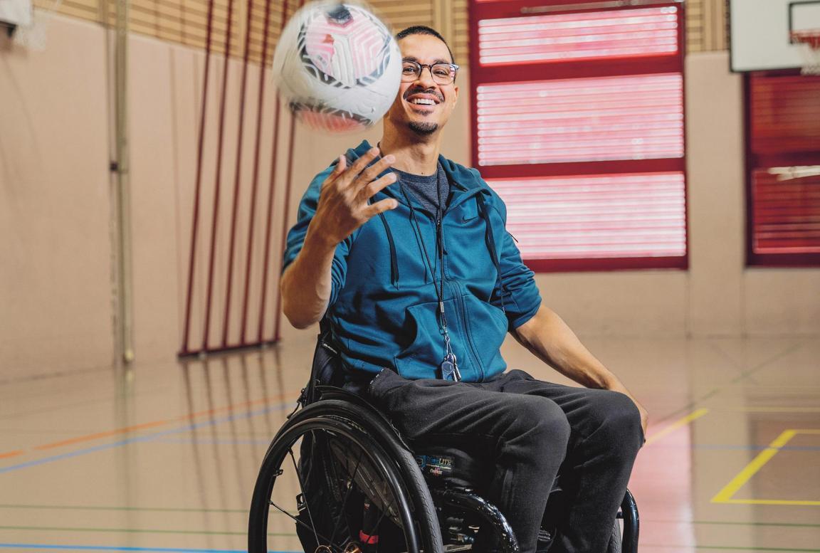Junger Mann im Rollstuhl in einer Turnhalle. Er dribbelt einen Fussball in der Luft