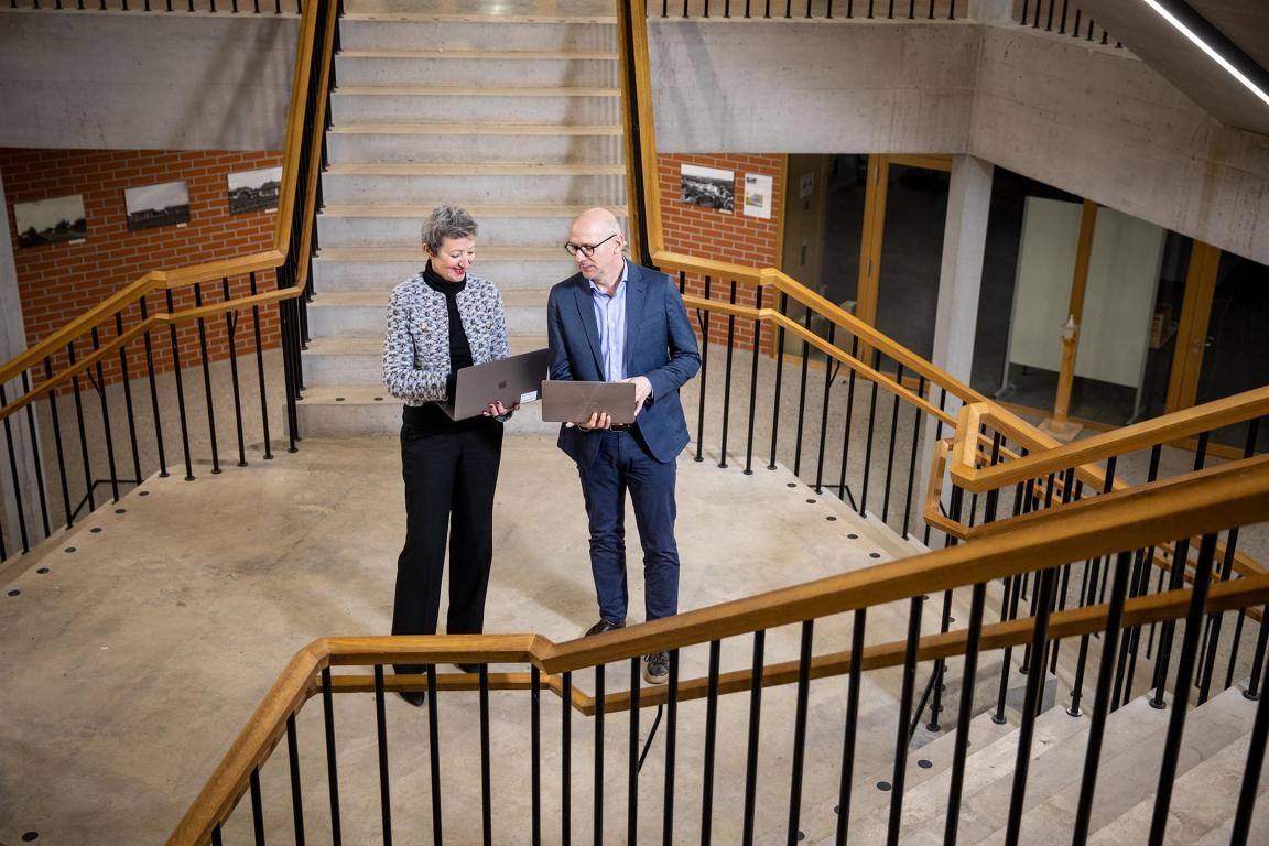 Ein Mann und eine Frau stehen auf der Treppe eines Schulhauses, beide mit einem PC in der Hand. 
