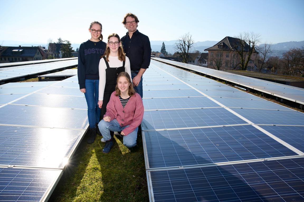 Drei Schülerinnen und ein Lehrer auf einem Flachdach mit Solarpanels. 