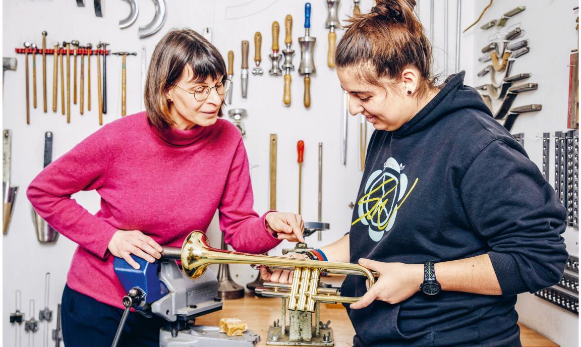 Eine Mitarbeitende und eine Lernende schauen sich in einer Werkstatt ein Blasinstrument an. 