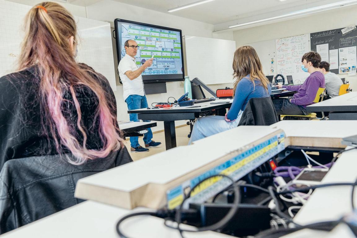 Zwei weibliche und zwei männliche Lernende sitzen in ihren Pulten in einem Klassenzimmer. Der Lehrer steht vor einer weissen Wandtafel und einem Bildschirm. 