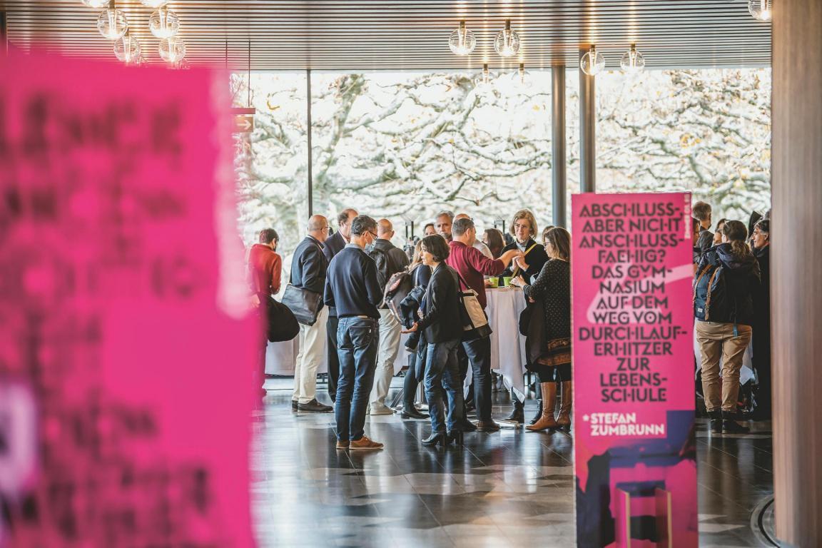 Verschiedene Personen haben sich in einem Foyer oder Saal zu einem Apéro zusammengefunden. Im Vordergrund stehen zwei Aufsteller mit Plakat.