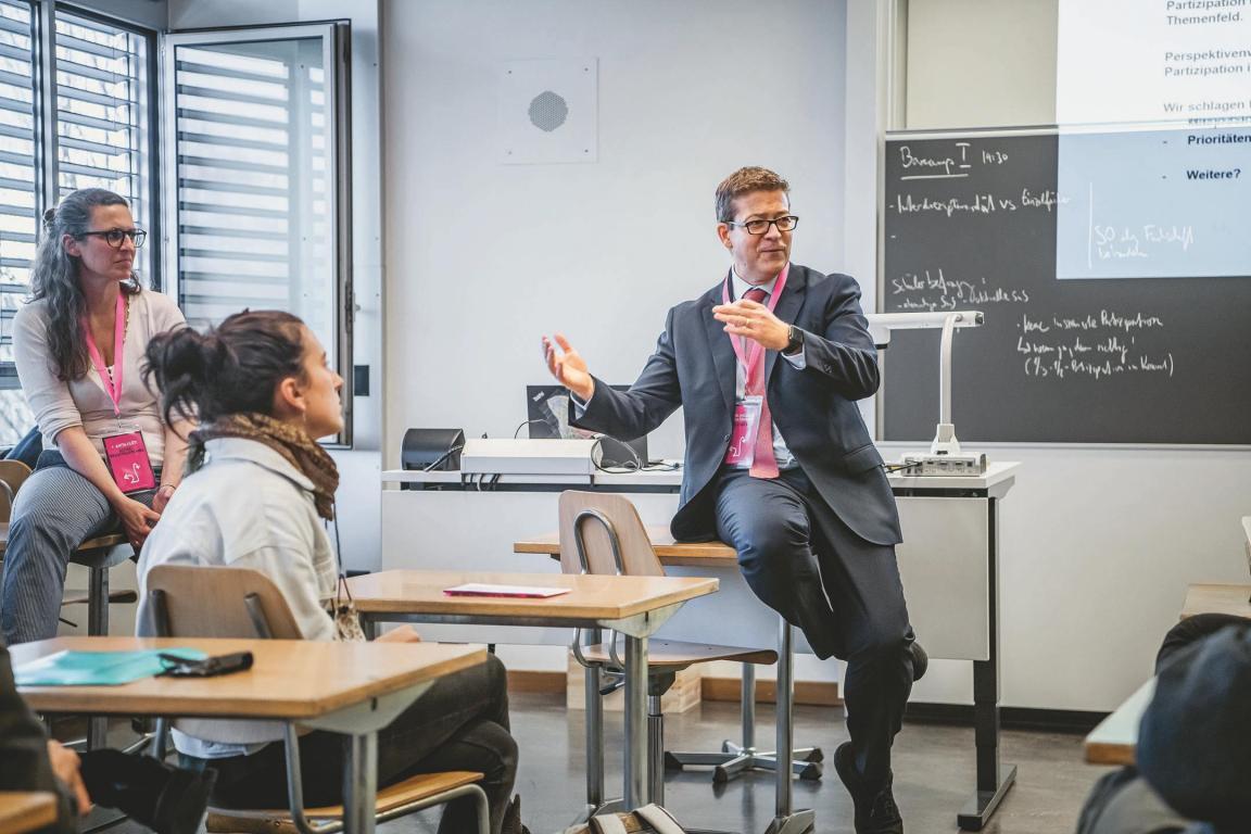 In einem Klassenzimmer hält Niklaus Schatzmann, Chef des Mittelschul- und Berufsbildungsamtes, einen Vortrag. Schatzmann sitzt auf einem Schulpult.
