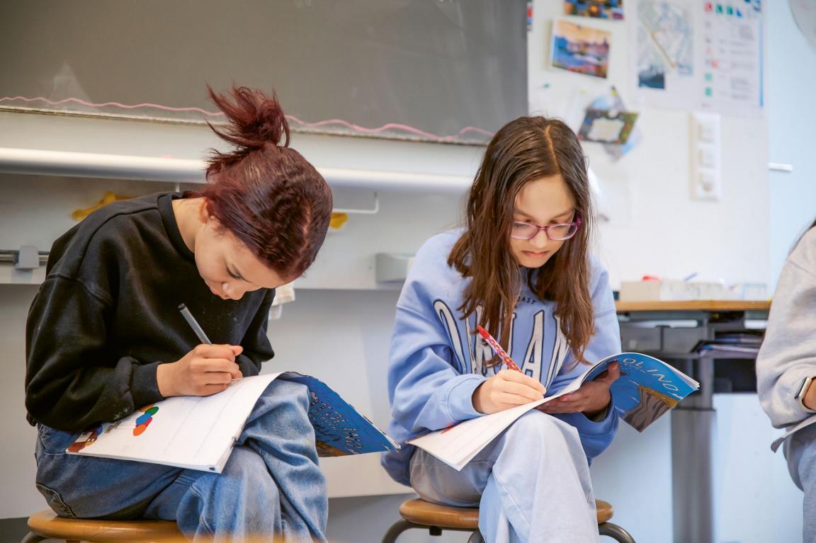 Zwei Schülerinnen sitzen auf Stühlen und haben ein Heft auf den Beinen. Darin notieren sie etwas. 