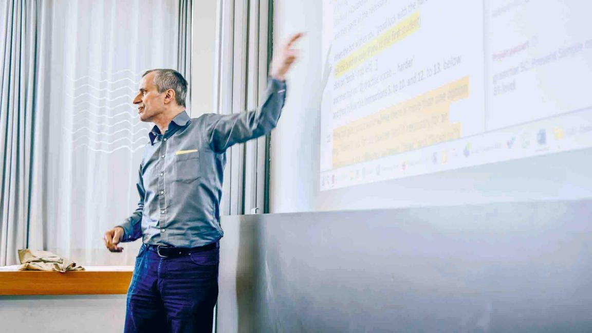 Michael Beusch im Schulzimmer, er zeigt auf  einen Text, der an die Wand projiziert ist. 