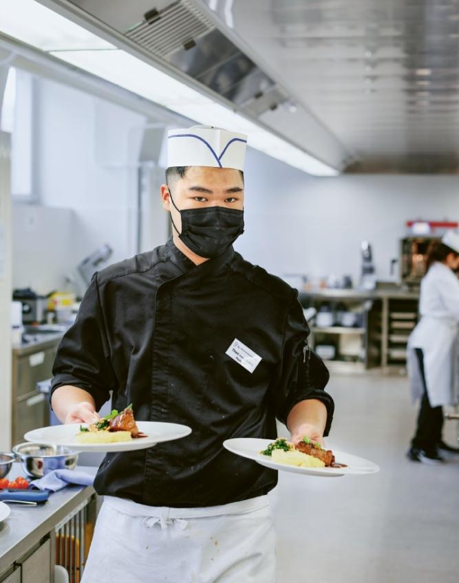 Ein junger Mann in Koch-Montur trägt zwei Teller mit schön angerichtetem Essen durch eine Grossküche