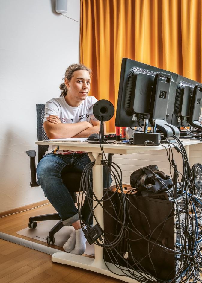 Ein junger Mann sitzt mit verschränkten Armen an einem Bürotisch. Auf dem Bürotisch stehen zwei Computermonitore sowie ein Mikrofon 