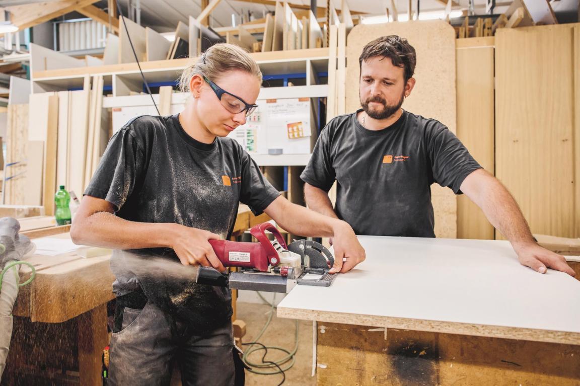 Der Ausbildner und die Lernende stehen in einer Schreinerei. Die Lernende sägt mit einer Maschine in ein Holzbrett