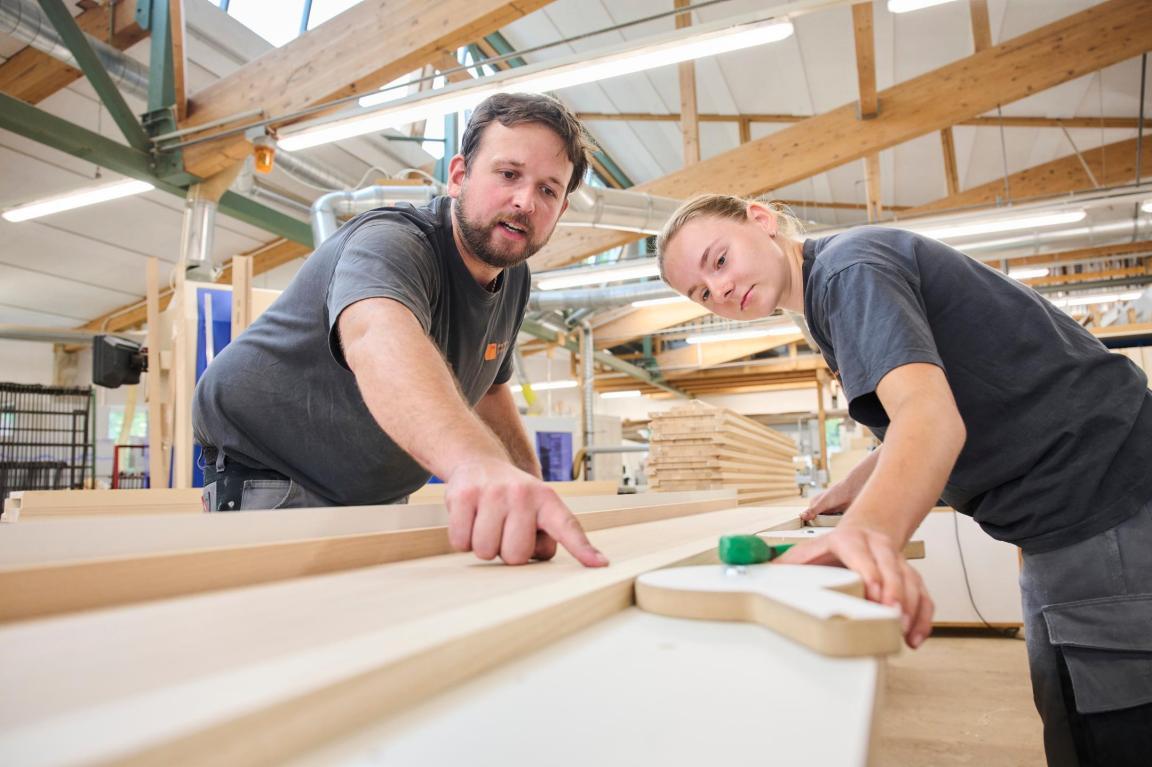 Der Ausbildner und die Lernende beugen sich über eine Holzlatte. Sie stehen in einer Schreinerei