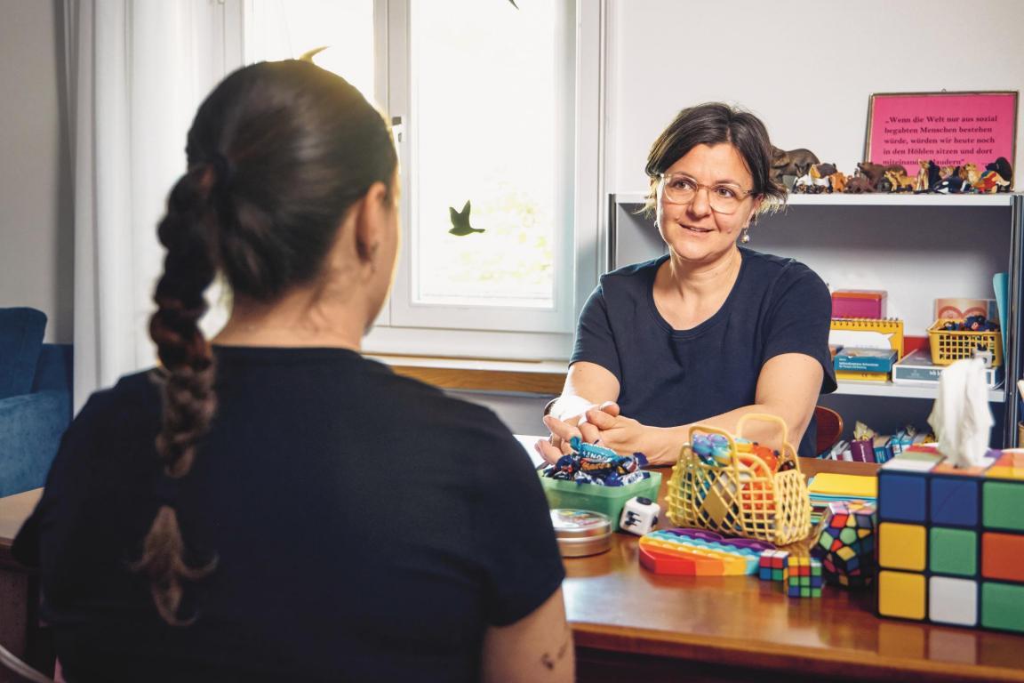  Jugendliche spricht mit der Schulsozialarbeiterin im Büro, auf dem Tisch stehen viele farbige Gegenstände. 