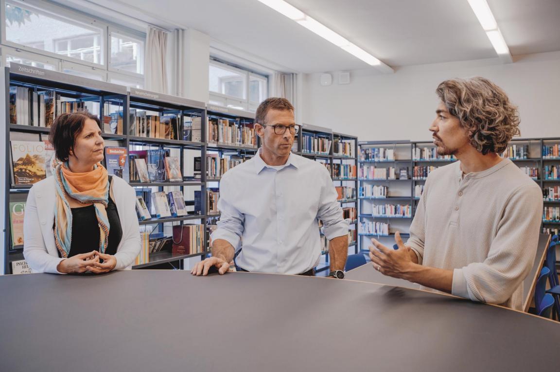 Zwei Männer und eine Frau stehen in einer Bibliothek um einen Tisch und sprechen  miteinander