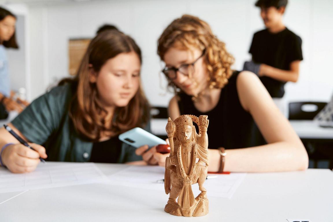 Im Vordergrund ist eine kleine Holzfigur zu sehen, die auf einem Schulpult steht. Im Hintergrund sitzen zwei Mädchen, die auf ein Smartphone blicken.