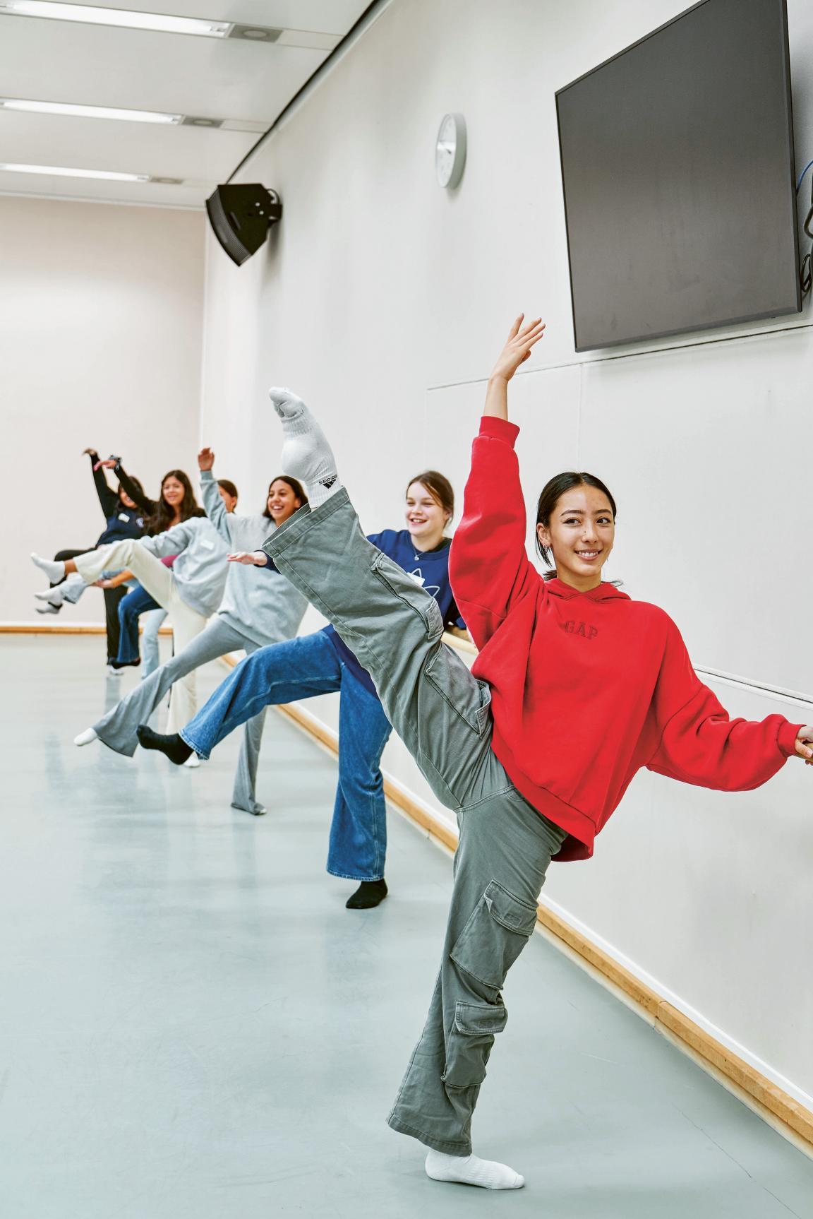 Schülerinnen sind an einer Ballettstange aufgereiht und heben einen Arm und einen Fuss in die Luft.