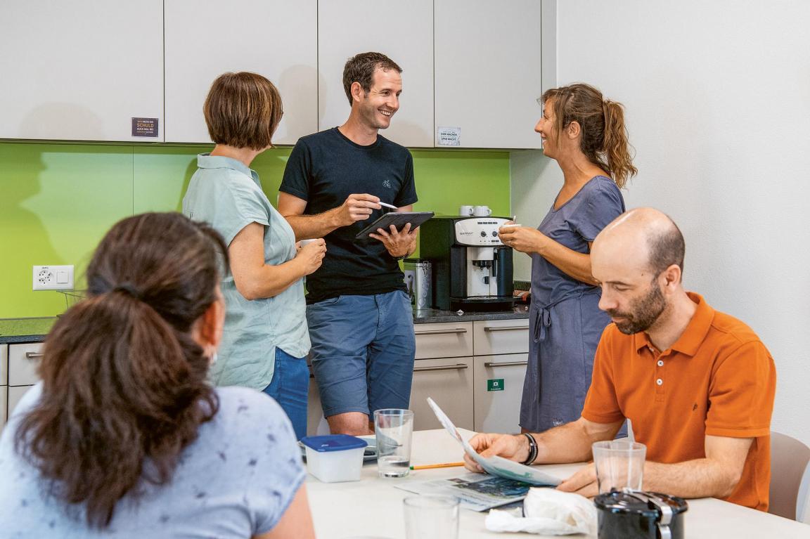 Drei Lehrpersonen unterhalten sich im Teamzimmer. Zwei Personen sitzen an einem Tisch und lesen Zeitung.