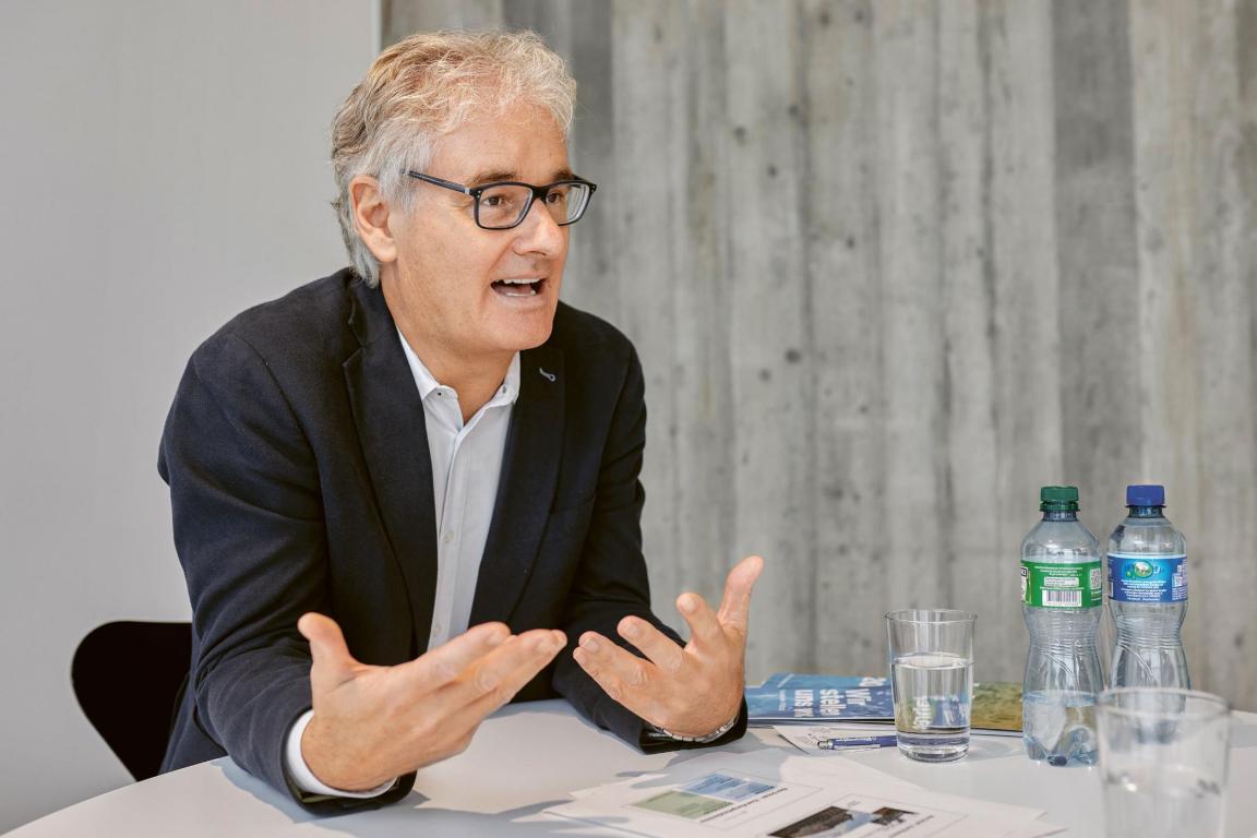 Patrick Heeb, Rektor des Berufsbildungszentrums Zürichsee, im Gespräch. Er gestikuliert mit den Händen. Vor ihm liegt eine Zeitung auf dem Tisch, daneben sind zwei Wasserflaschen zu sehen.