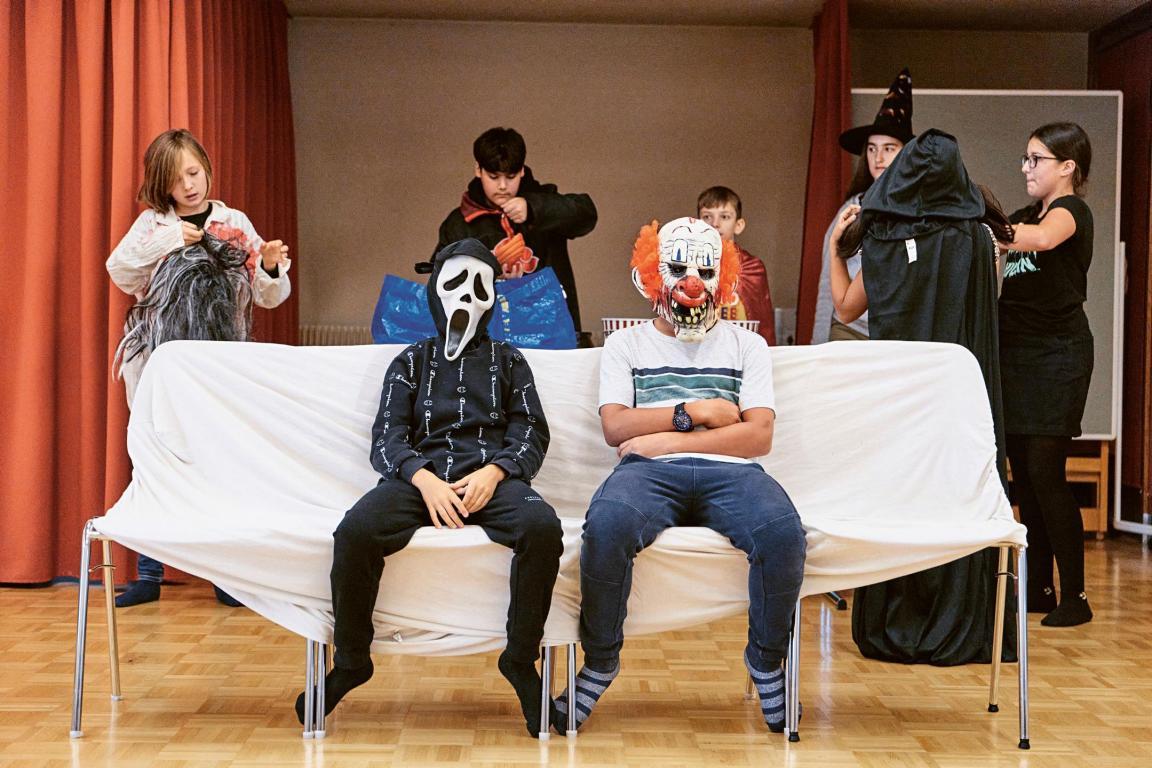 Schüler der Primarschule Oberengstringen bereiten sich auf die Aufführung des Theaterstücks vor, das sie selber entwickelt haben. Im Vordergrund sitzen bereits zwei von ihnen auf Stühlen, die mit einem Leintuch verhüllt sind. Sie haben beide eine Maske auf: Der Junge links trägt den «Schrei» von Edvard Munch. Der Junge rechts trägt eine etwas gruselige Clown-Maske.
