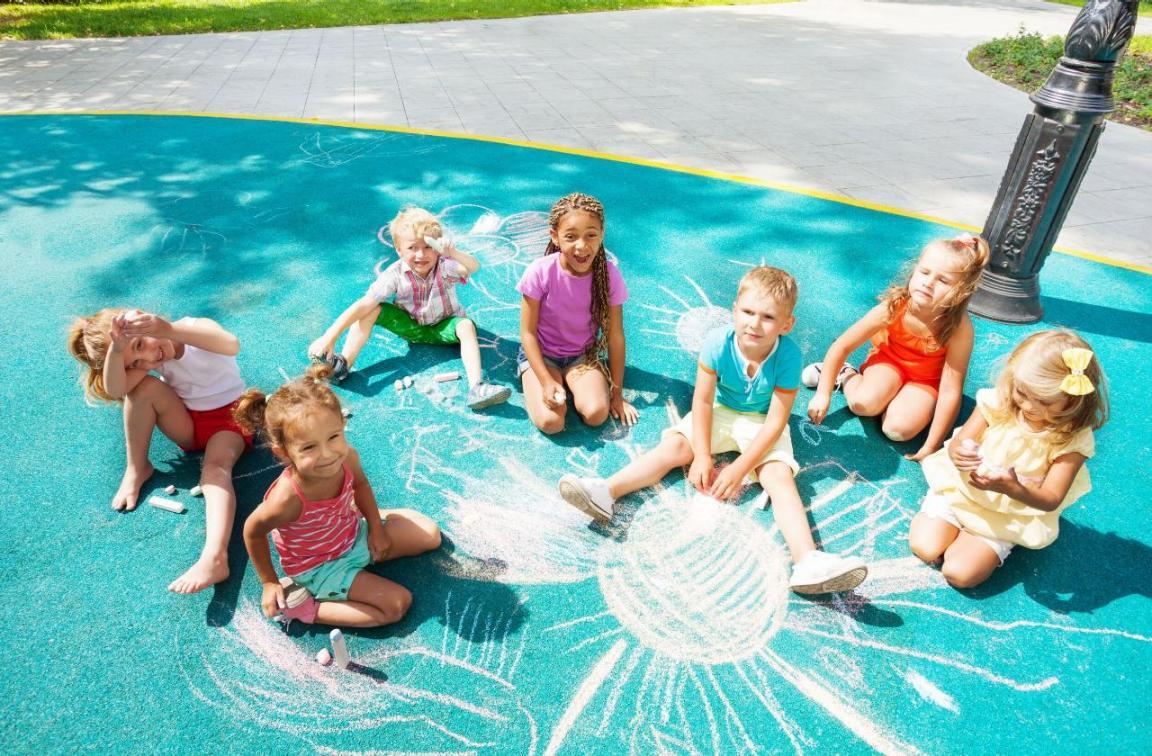 Sieben Kindern sitzen bei Sonnenschein auf einem türkis bemalten Asphalt und malen mit Kreide