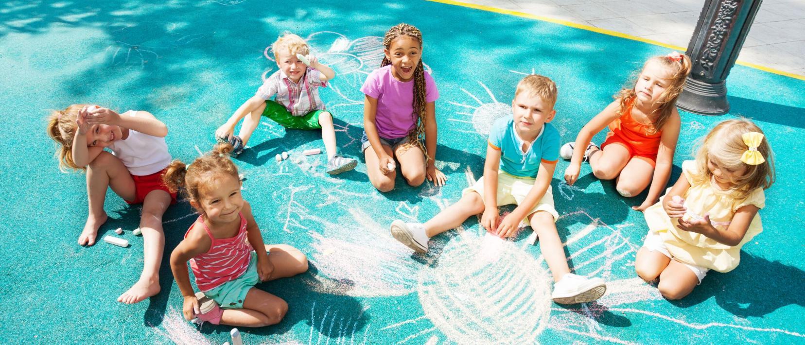 Sieben Kindern sitzen bei Sonnenschein auf einem türkis bemalten Asphalt und malen mit Kreide. Die Kinder sind bunt gekleidet und sehen fröhlich aus.