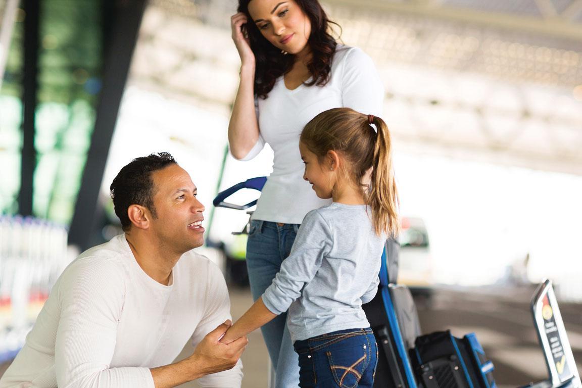 Vor einem Flughafeneingang kniet ein Mann vor einem kleinen Mädchen und hält ihre Hand, während eine Frau mit Gepäckwagen stehend auf die beiden herunterschaut.