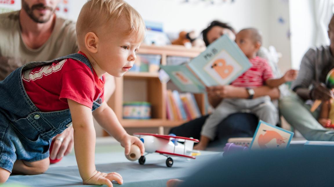 Ein Kleinkind spielt mit einem Spielzeugflugzeug, während Eltern mit Kindern Bilderbücher anschauen.