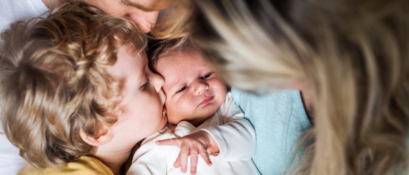 Ein Kleinkind küsst ein Baby auf die Wange, während beide von ihren Eltern liebevoll gehalten werden.
