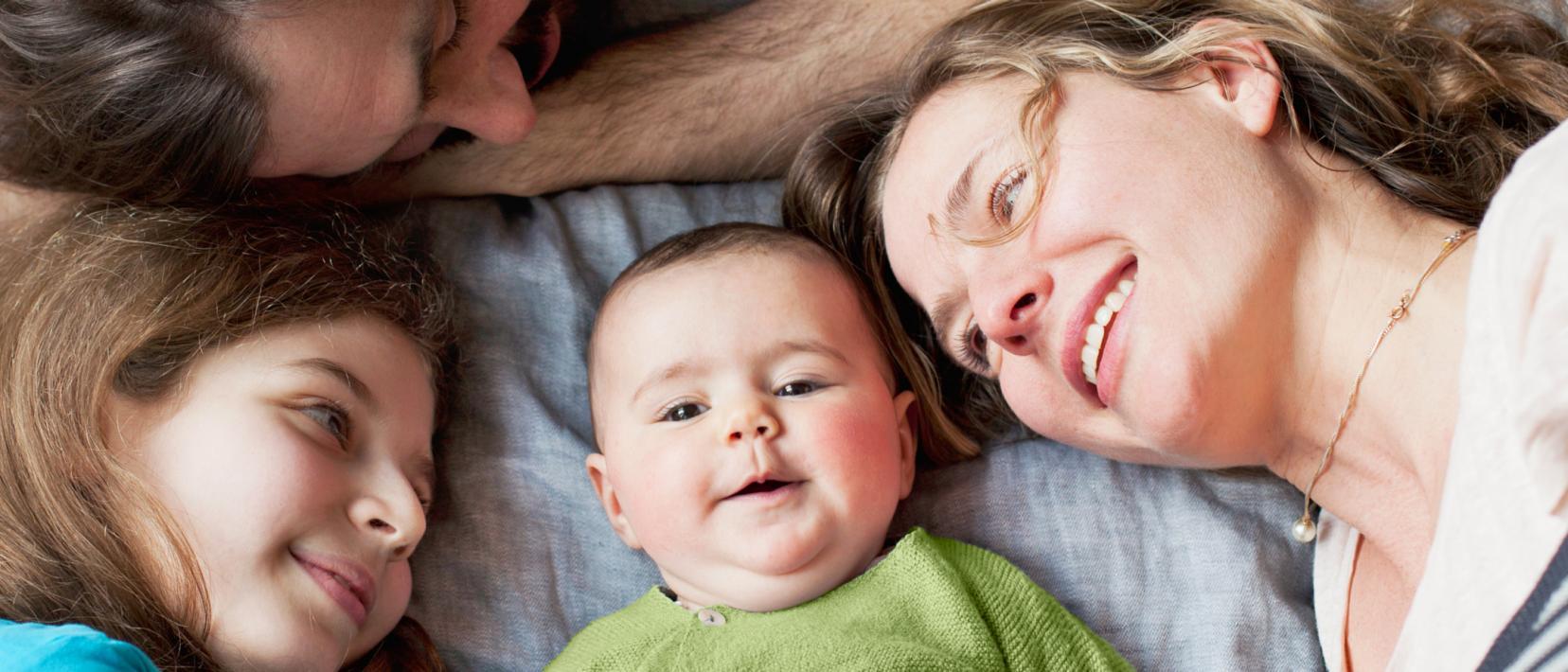 Eine Frau, ein Mann, ein Baby und ein Mädchen liegen auf einem grauen Tuch, halten die Köpfe zusammen und werden von oben fotografiert.