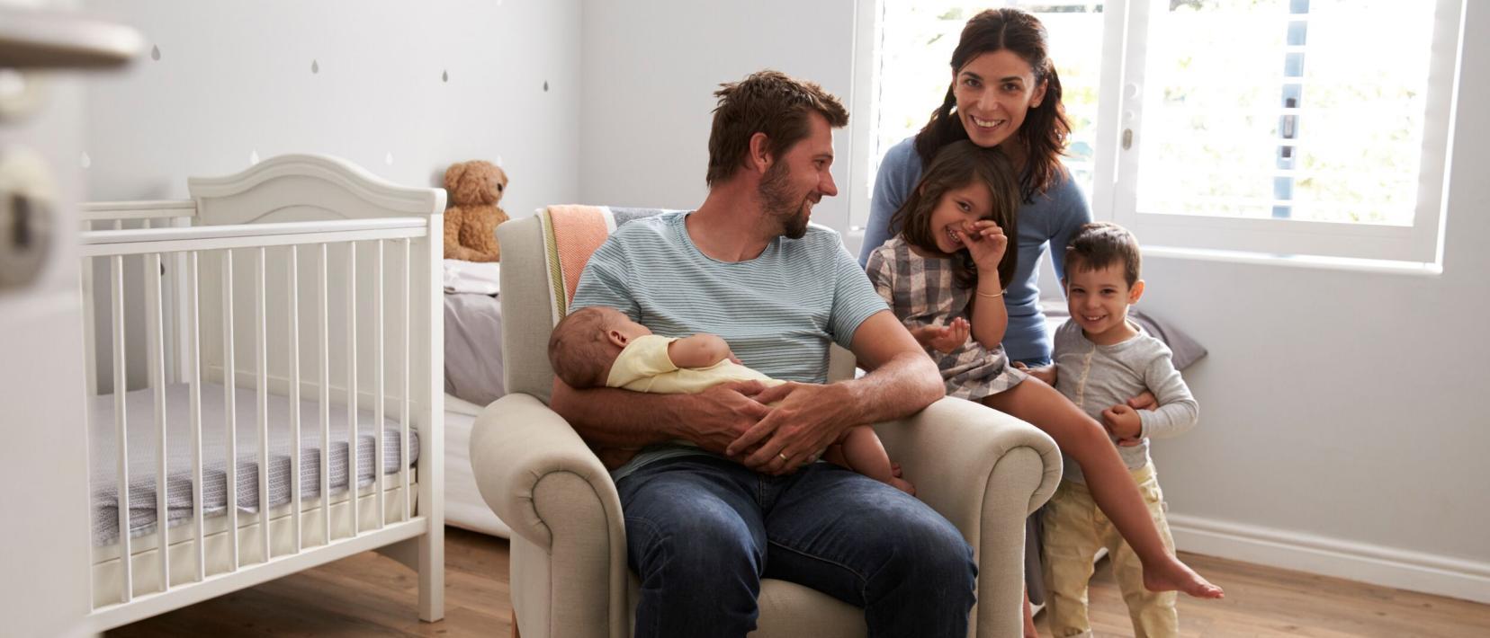 In einem hellen Raum mit Babybett und Fenster sitzt ein Mann auf einem Stuhl mit einem Baby im Arm und neben ihm hält eine Frau zwei Kleinkinder im Arm.