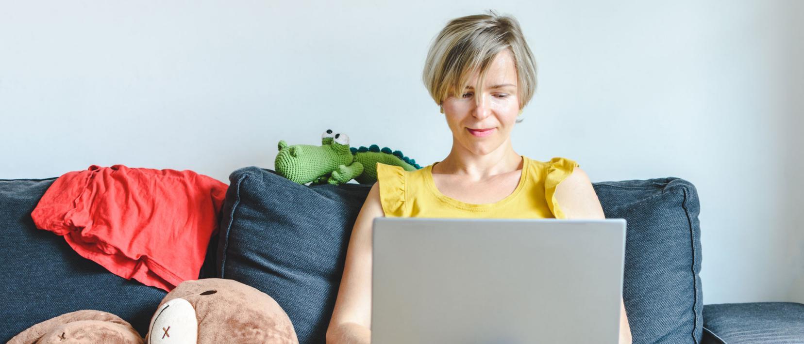 Blonde Mutter sitzt auf dem Sofa mit dem Laptop vor sich.