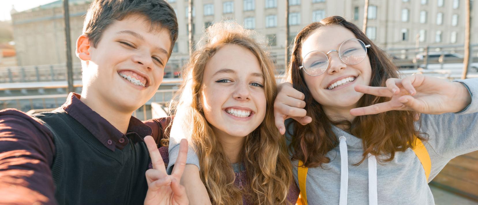 Ein Junge und zwei Mädchen im Teenie-Alter machen lachend und Victory-Zeichen zeigend in der Stadt ein Selfie von sich.