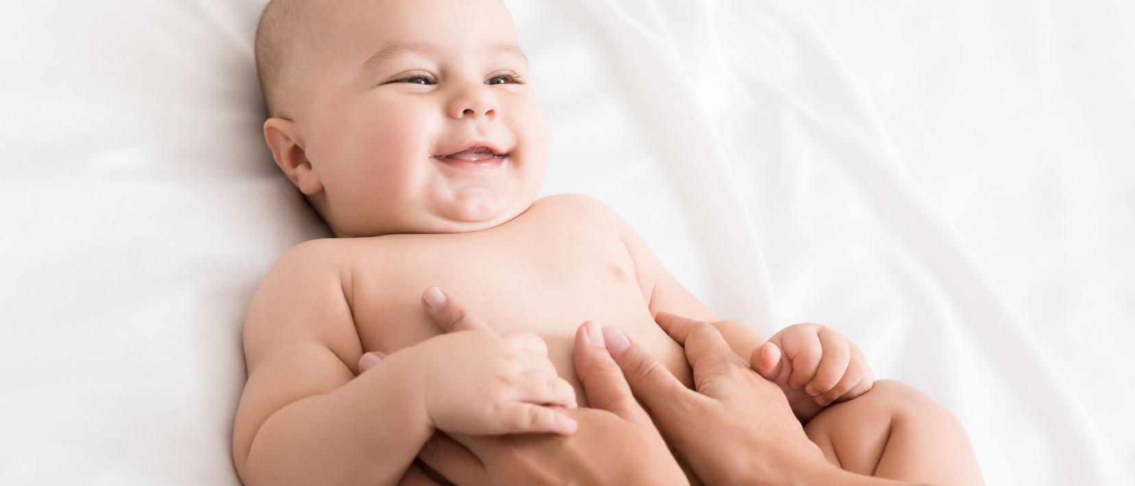 Ein lächelndes Baby geniesst die Bauchmassage von Frauenhänden.