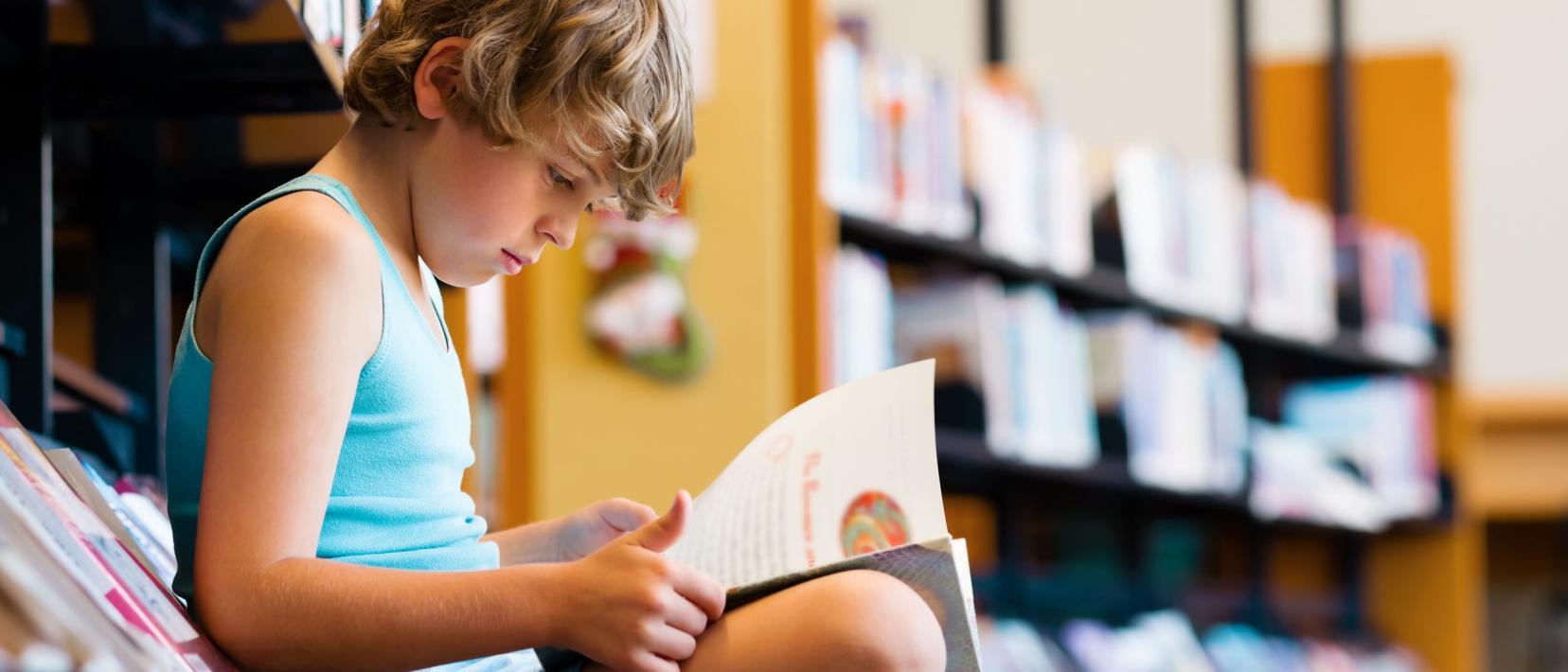 Ein Junge sitzt in einer Bibliothek vor einem Bücherregal und blättert in einem Buch.
