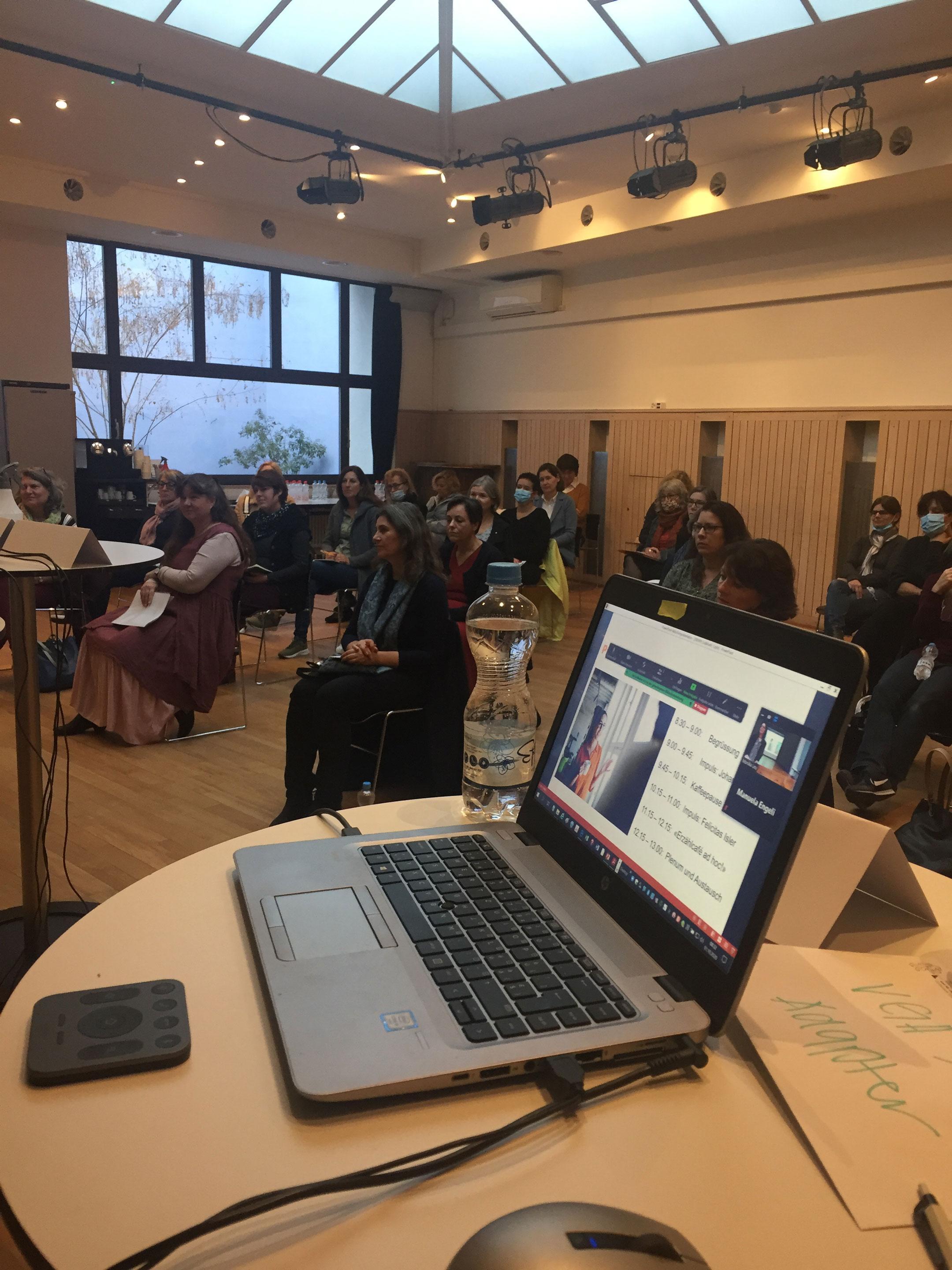 In einem Saal steht im Vordergrund auf einem runden Stehtisch ein geöffnetes Laptop. Im Hintergrund sieht man ein mehrheitlich weibliches Publikum.