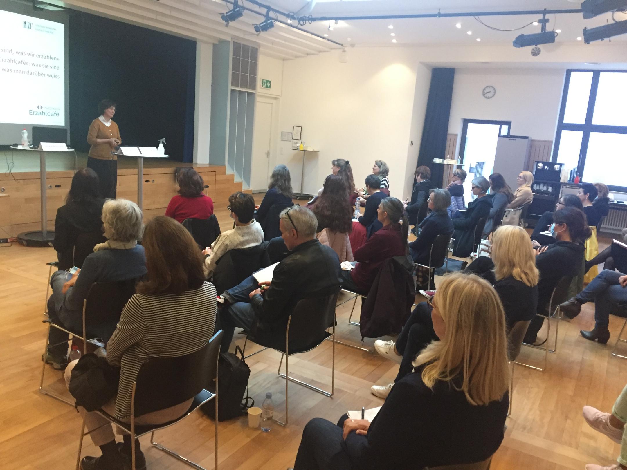 Ein interessiertes Publikum hört einer Rednerin zu die vor einer Projektionswand steht.