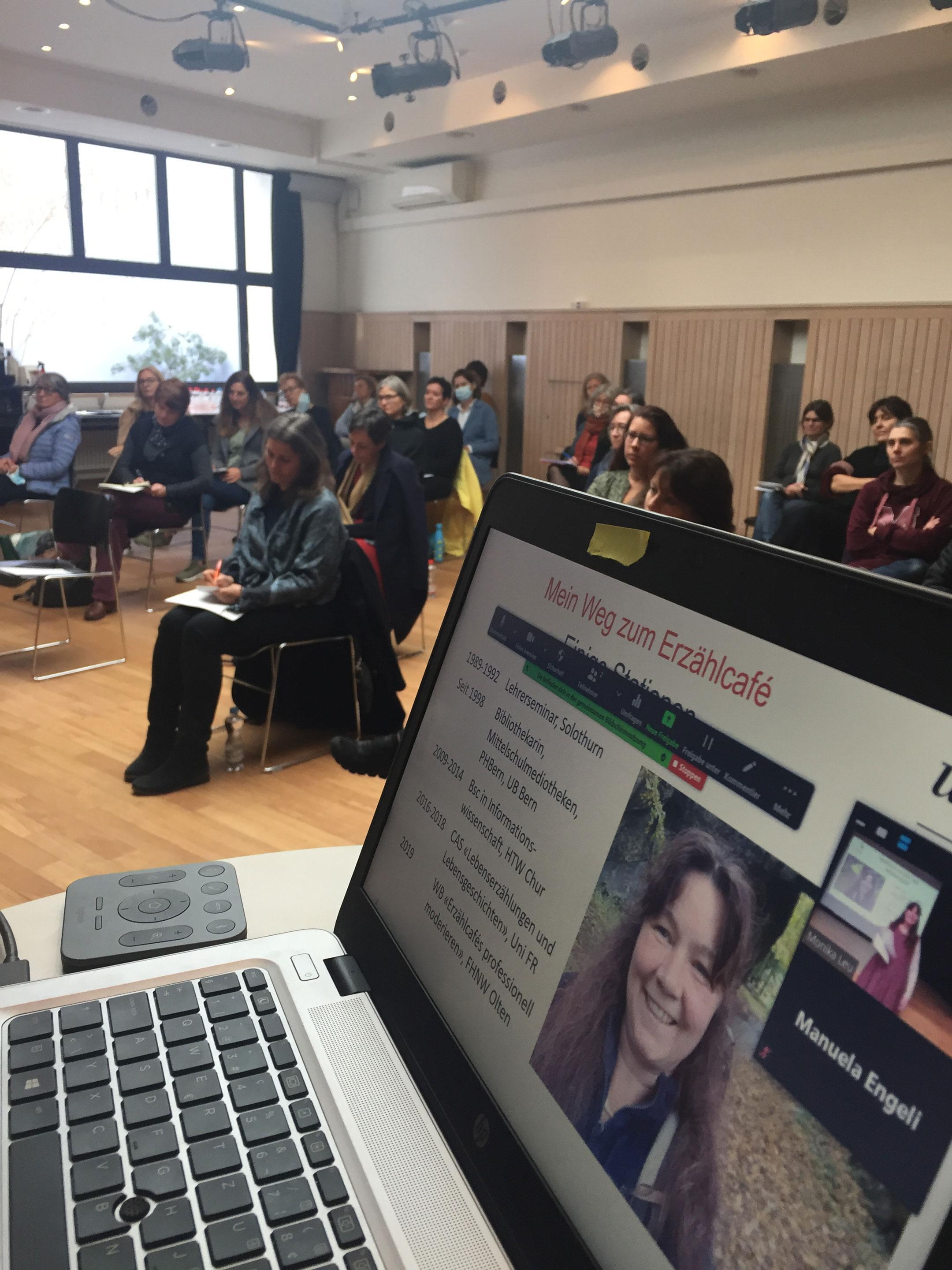 In einem Saal sieht man auf einem geöffneten Laptop eine Seite der Präsentation die gerade gezeigt wird. Im Hintergrund sieht man die Teilnehmenden sitzen.