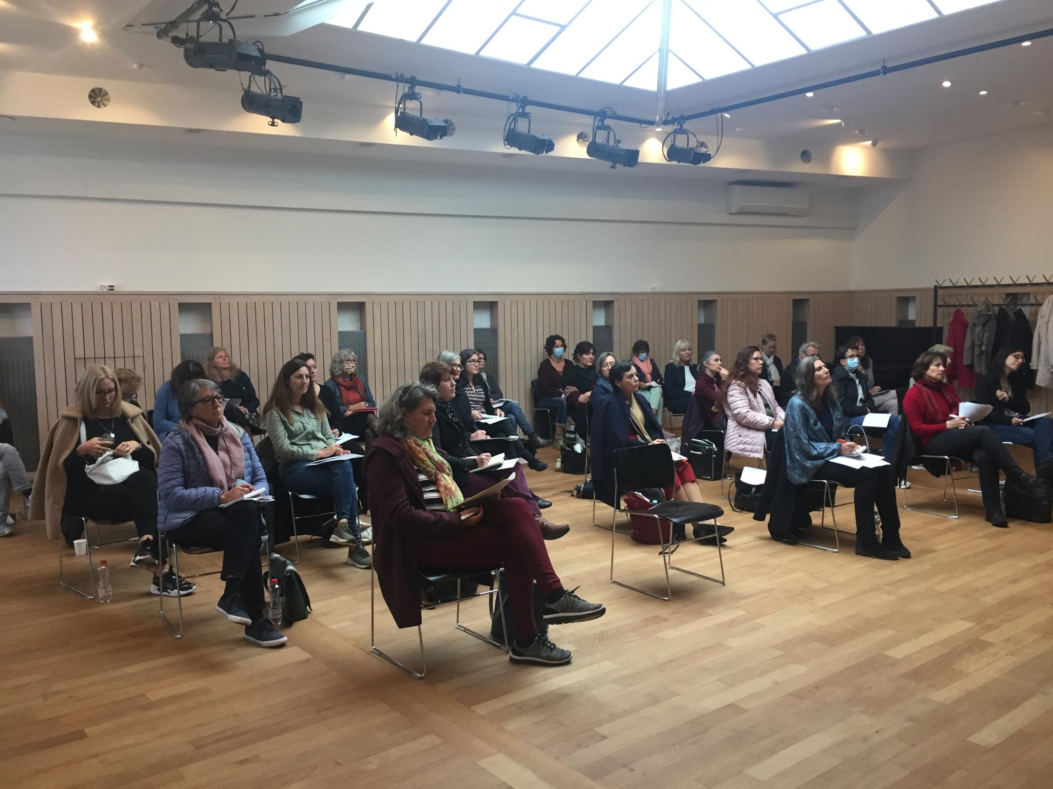In einem Saal mit Oberlicht sitzt das Publikum mit genügendem Abstand zueinander und sieht nach vorn.