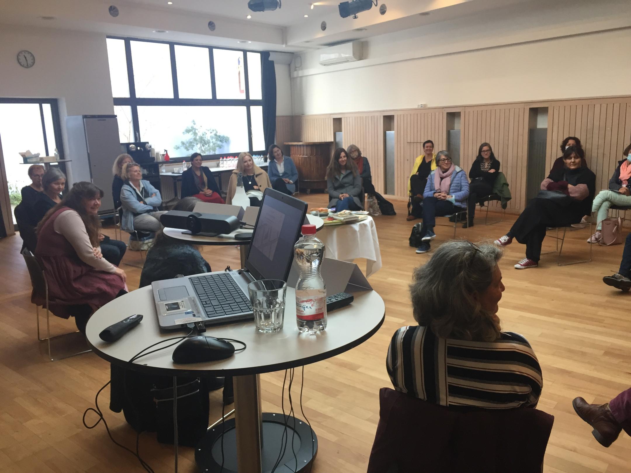 In einem Saal steht im Vordergrund auf einem runden Stehtisch ein geöffnetes Laptop. Frauen sitzen in einer Disskusionsrunde drum herum.