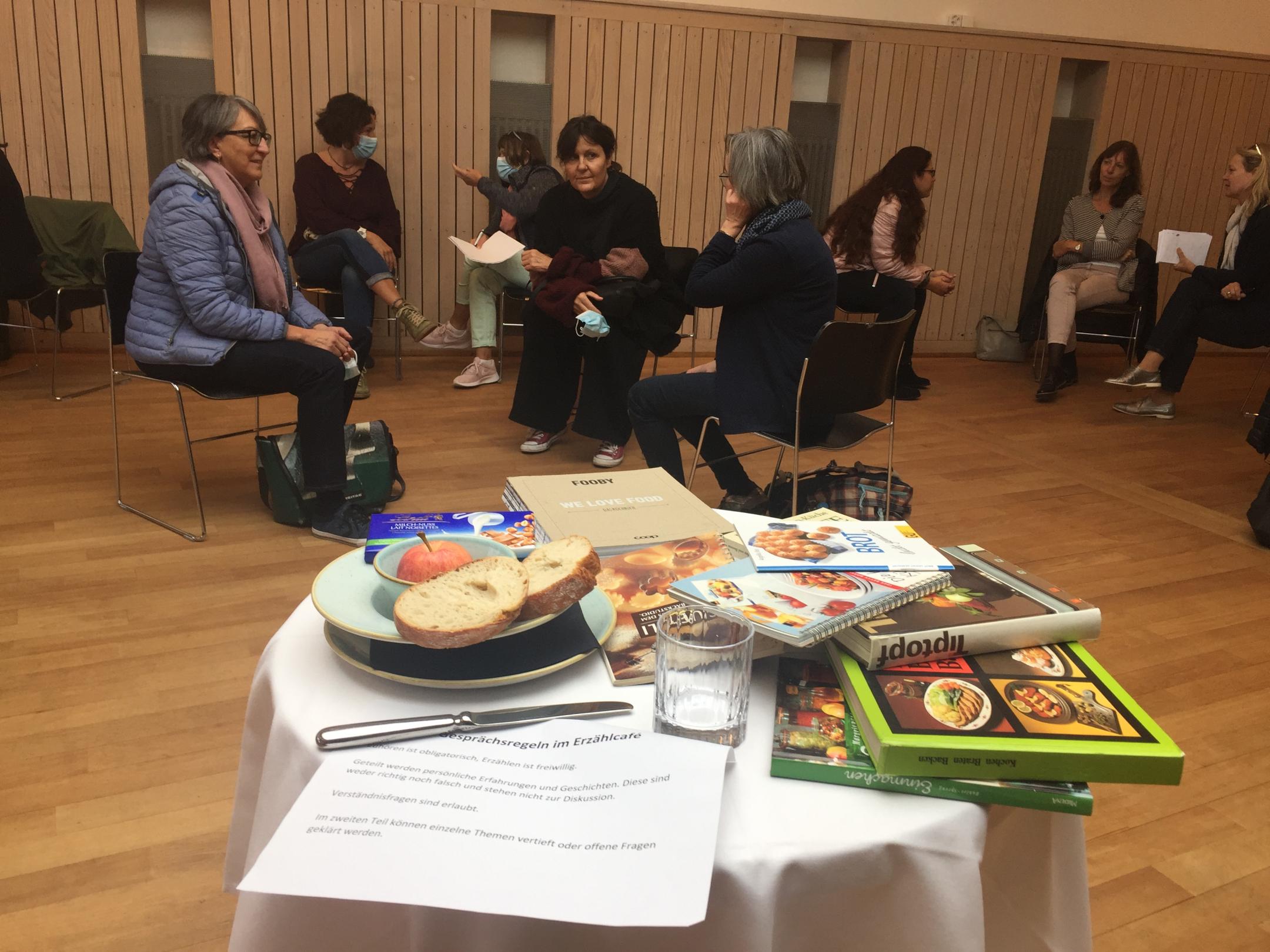 Im Vordergrund steht ein runder Tisch, der mit einem weissen Tischtuch gedeckt ist. Darauf liegen mehrere Kochbücher, ein Teller mit zwei Scheiben Brot einem Apfel und einer Tafel Schokolade, ein Messer und ein Wasserglas. Im Hintergrund diskutieren Frauen in Dreier-Gruppen.