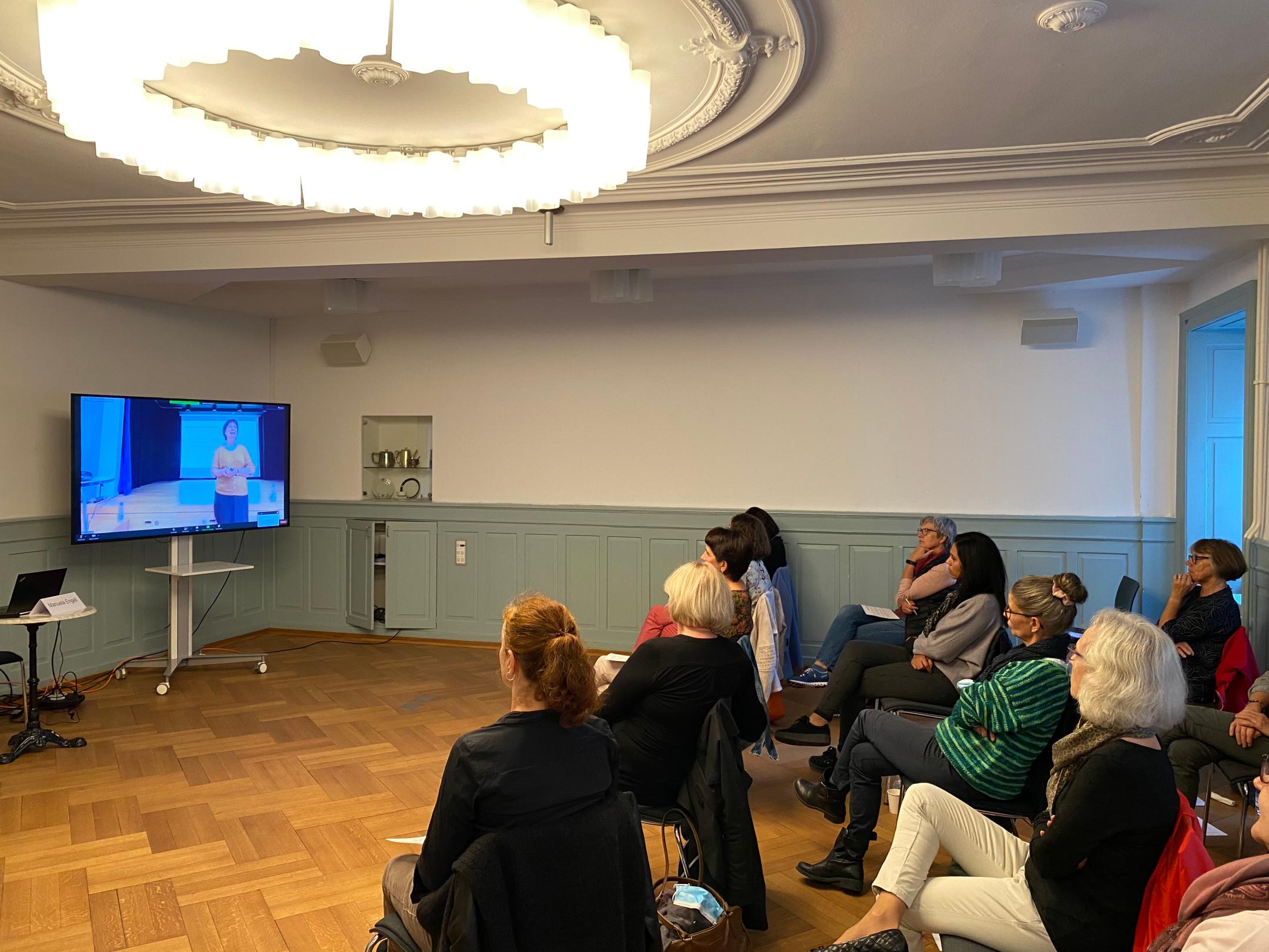 In einem kleineren Saal, mit schöner Stuckdecke und einer grossen runden Lichtquelle, hört ein interessiertes Publikum der Rednerin zu die über einen Screen übertragen wird.