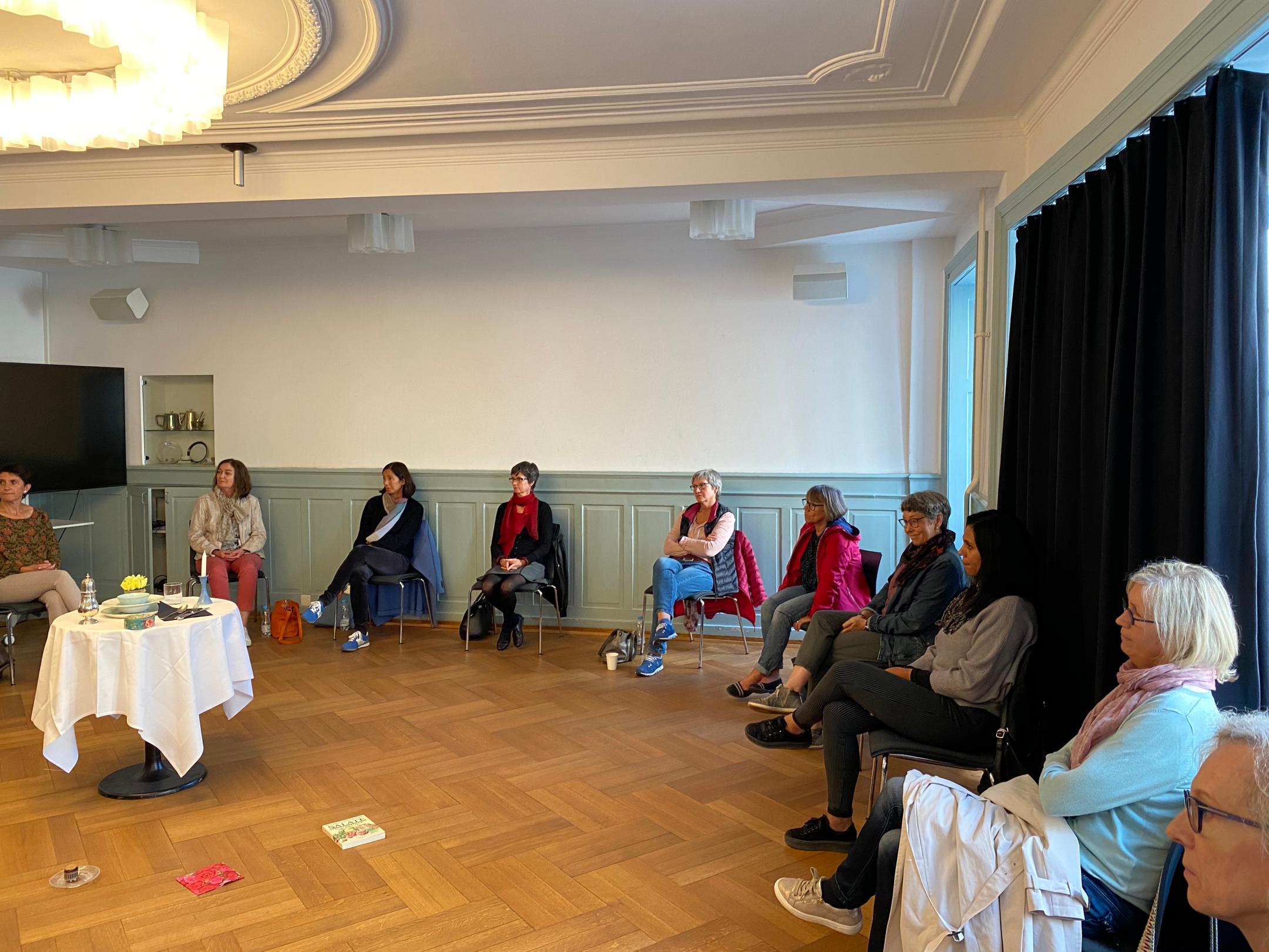Frauen sitzen in einem Raum an den Wänden entlang. Rechts ist ein schwarzer Vorhang vor einem grossen Fenster gezogen. Links steht ein gedeckter Tisch vor dem auf dem Parkett ein Buch, eine Serviette und ein Dessert auf einem Teller liegen.