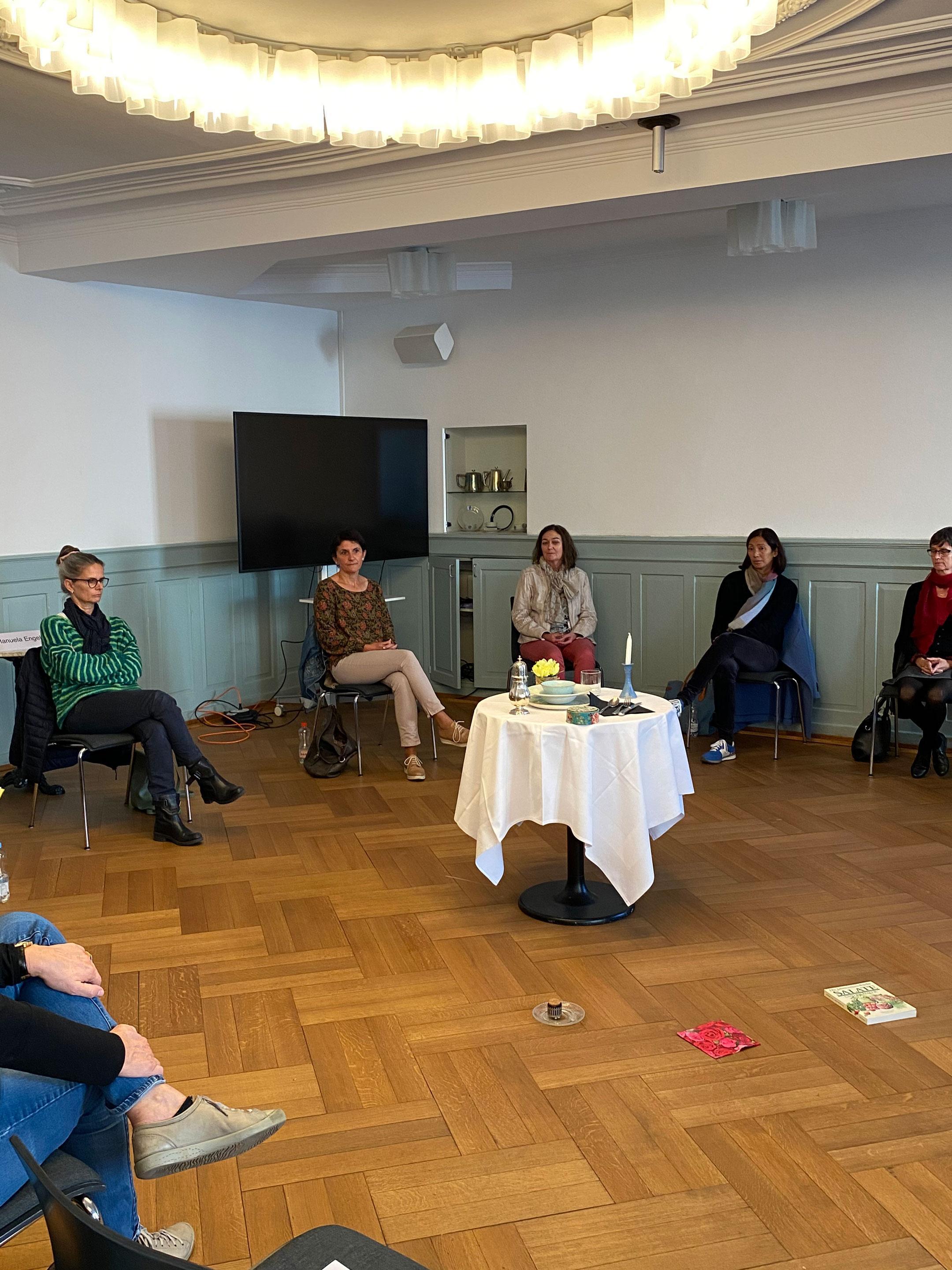 Frauen sitzen in einem Raum an den Wänden entlang. Rechts ist ein schwarzer Vorhang vor einem grossen Fenster gezogen. In der Mitte steht ein gedeckter Tisch vor dem auf dem Parkett ein Buch, eine Serviette und ein Dessert auf einem Teller liegen.