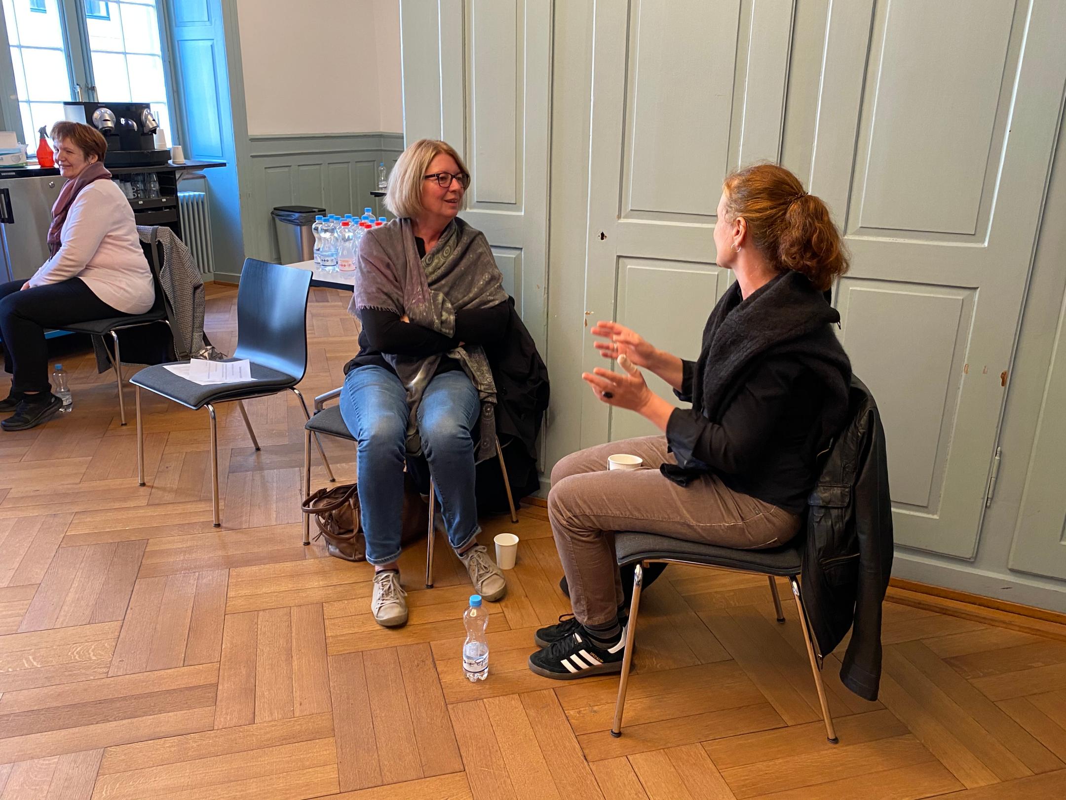 Zwei Frauen diskutieren gestenreich miteinander. Links von ihnen steht vor einem Fenster eine Kaffemaschine vor der eine weitere Frau sitzt.