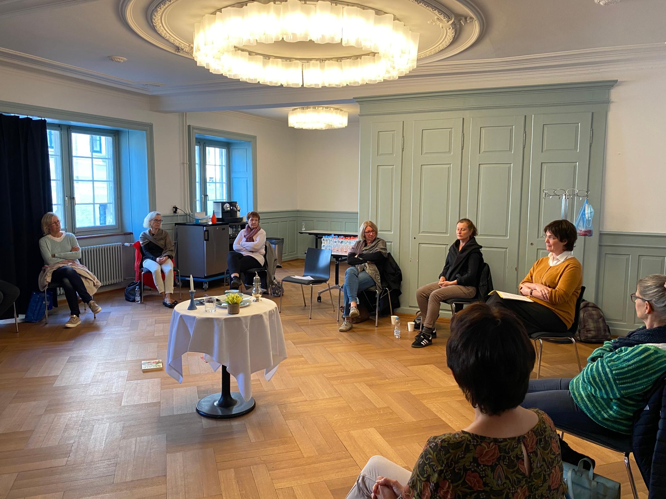 Frauen sitzen in einem grossen Kreis um einen weiss gedeckten Tisch und diskutieren miteinander.