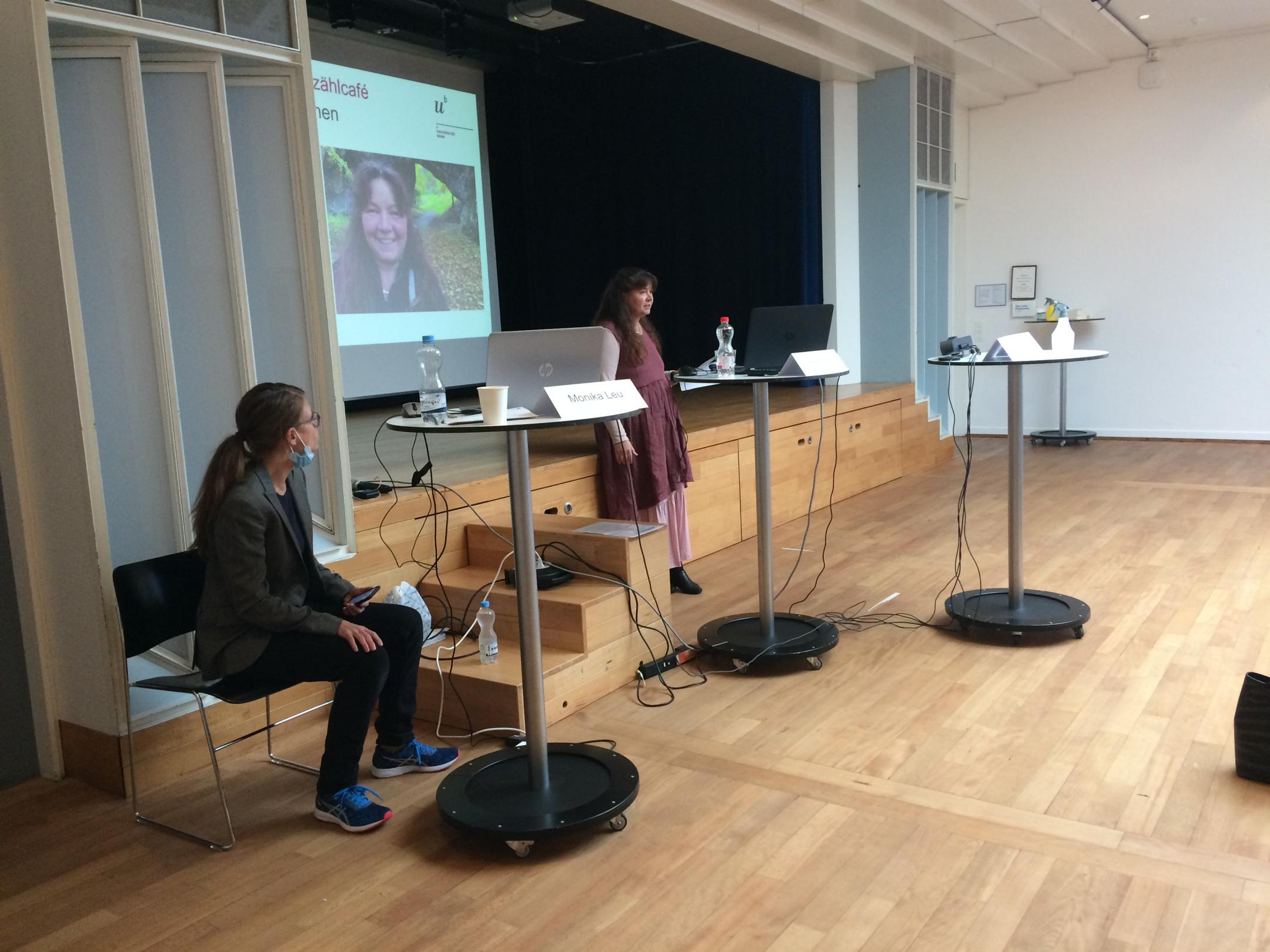 Eine Rednerin steht vor der Bühne auf der die Präsentation projiziert wird. Die Moderatorin sitzt links von ihr.