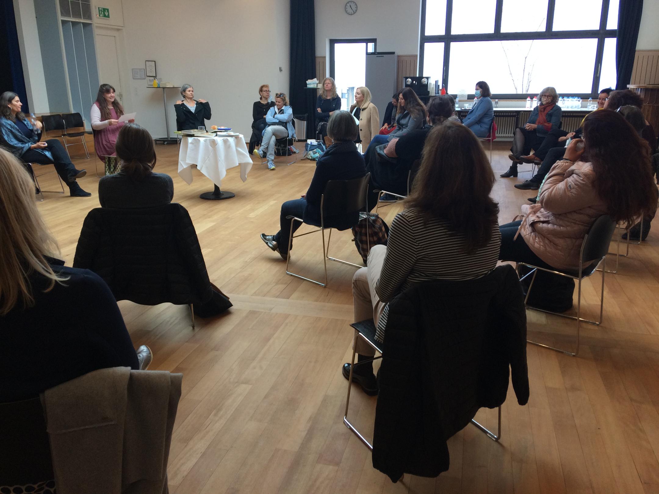 Frauen sitzen in einem grossen Kreis um einen weiss gedeckten Tisch und hören der Rednerin zu. Zum Teil tragen die Frauen Schutzmasken.