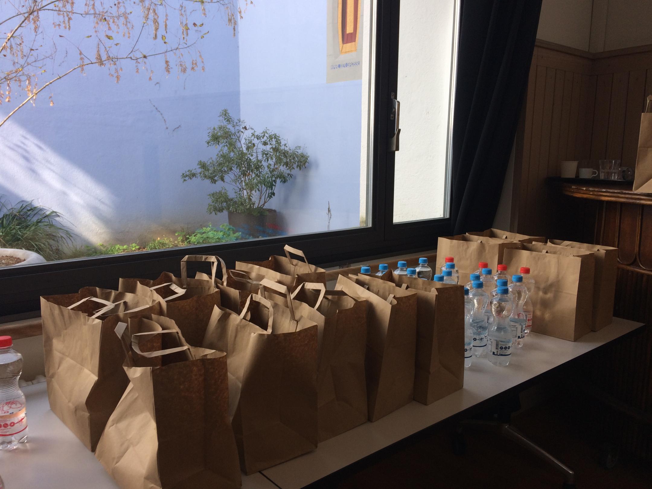 Vor einem grossen Fenster stehen auf Tischen braune Papiersäcke und kleine PET-Flachen mit Mineralwasser.