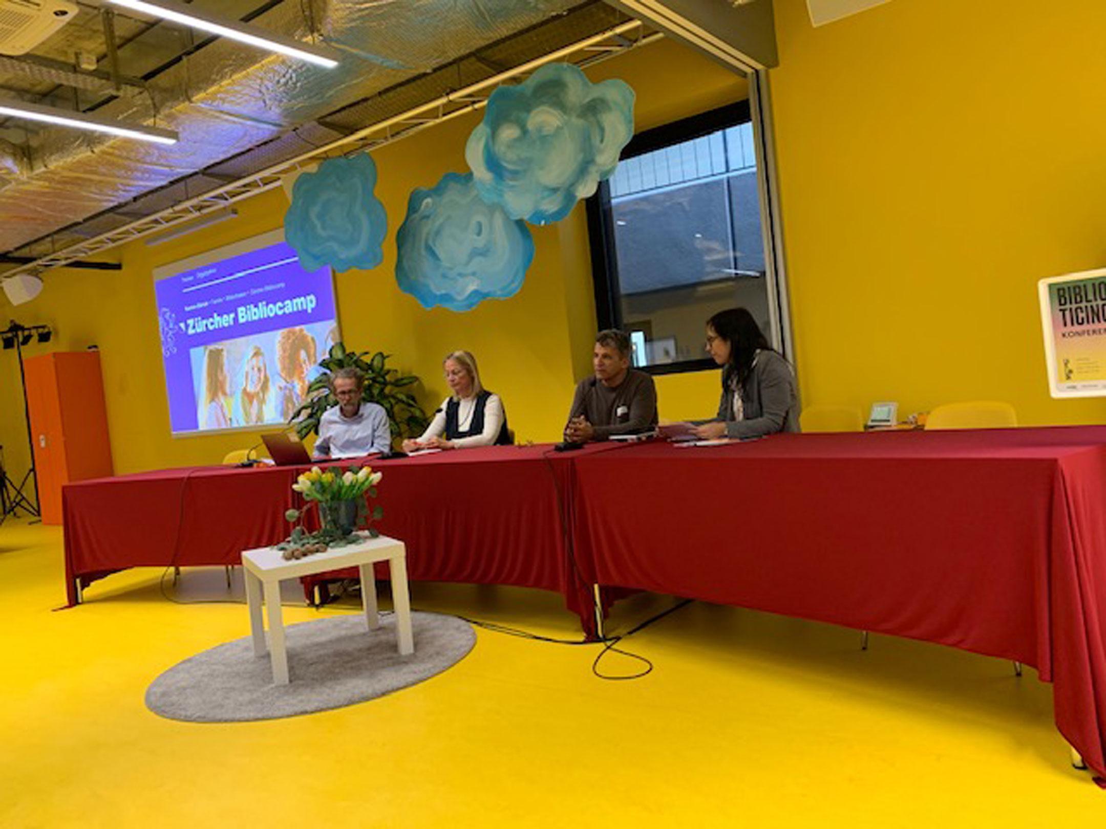 Die vier Moderierenden sitzen vor einer gelben Wand an einem Tisch.