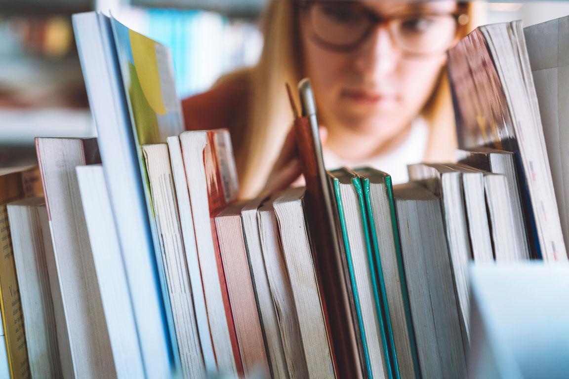 Junge blonde Frau mit Brille, sucht in der Bibliothek nach Büchern. 
