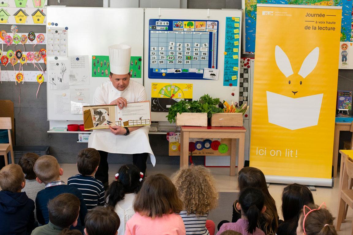 Ein als Koch verkleideter Mann liest einer Schar kleiner Kinder aus einem Buch vor.