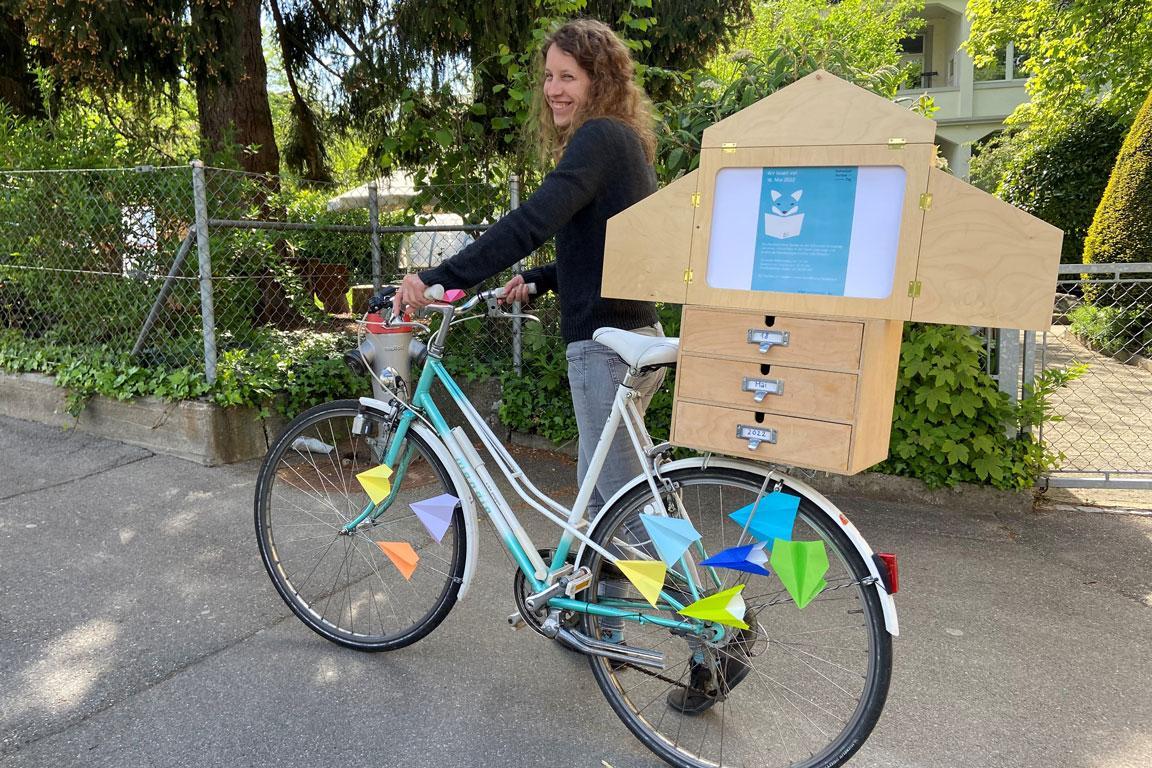 Eine Frau schiebt ein Fahrrad mit einer Kamishibai auf dem Gepäckträger.